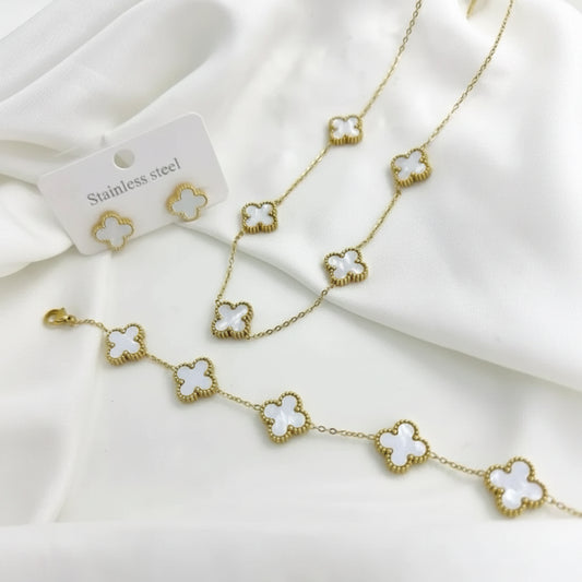 Gold-tone jewelry set featuring a necklace, bracelet, and matching stud earrings with white clover motifs, displayed on a white fabric background with a small card labeled 'Stainless steel'.
