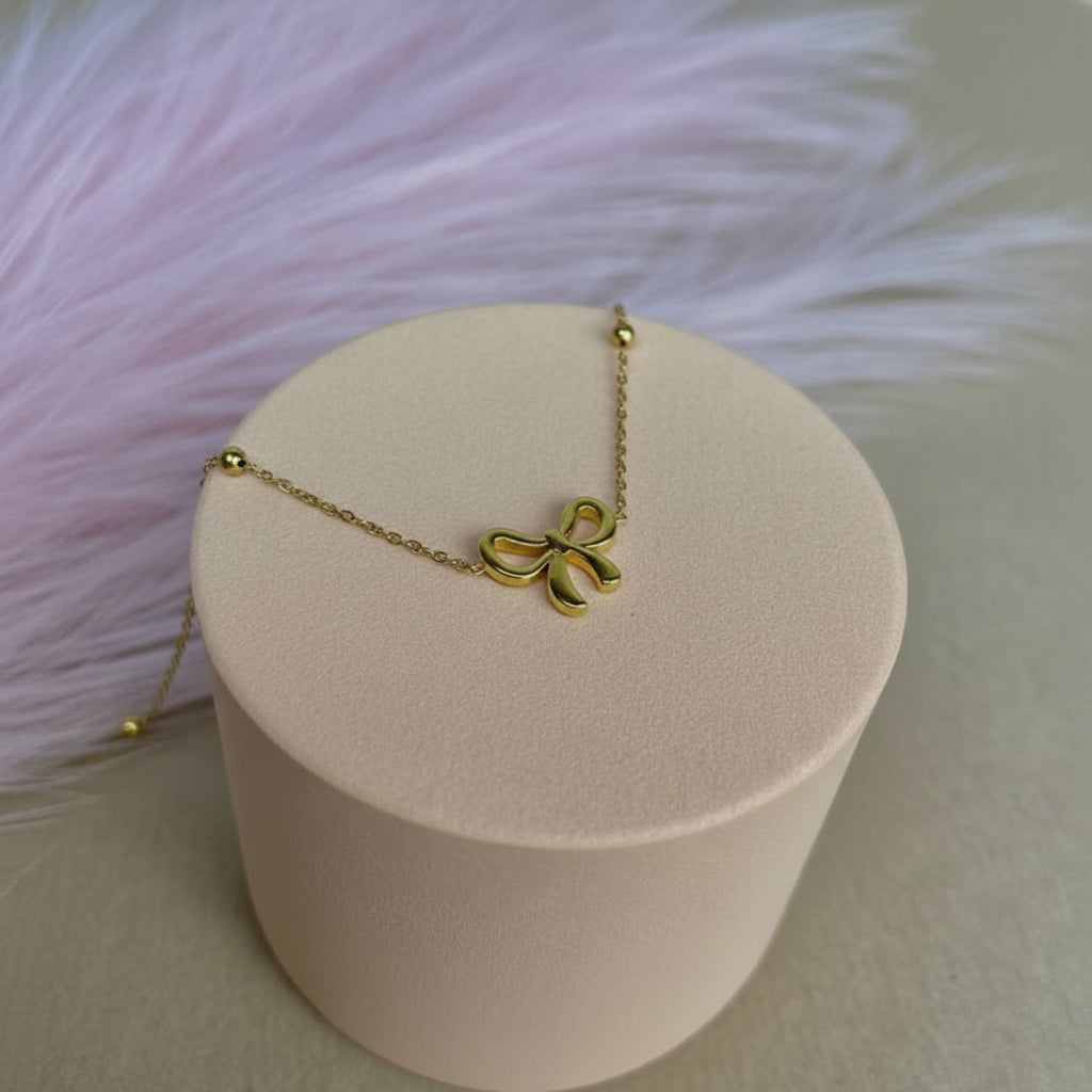 A delicate gold chain necklace with a small bow-shaped pendant, displayed on a round beige jewelry box against a soft pink fluffy background.