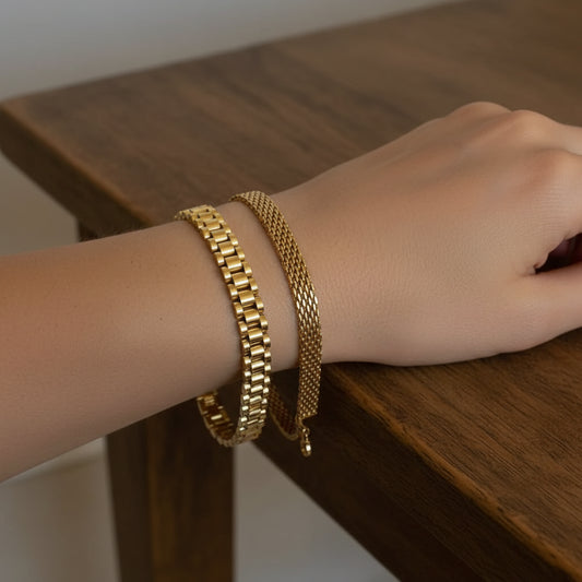Two gold-tone metal chains, one with a polished rectangular link design and the other with a fine mesh pattern, draped over a woven beige chair.