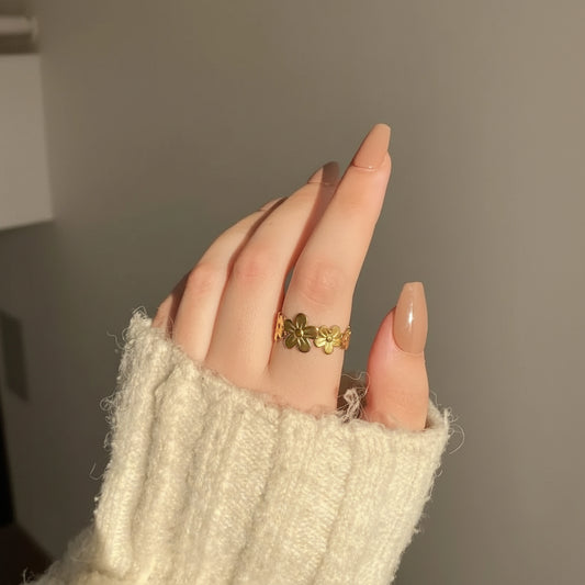 A close-up of a hand wearing a gold ring with three small flower motifs, styled with a cream-colored knit sweater sleeve.