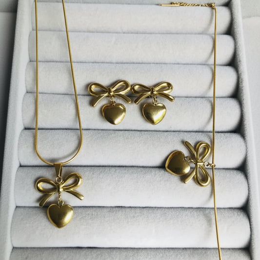 Gold jewelry set featuring a necklace, earrings, and ring with bow and heart pendants, displayed on a light gray velvet jewelry tray.