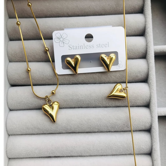 Gold heart jewelry set including a necklace, stud earrings, and a heart-shaped pendant, displayed on a gray jewelry tray with a white card labeled 'Stainless steel'.