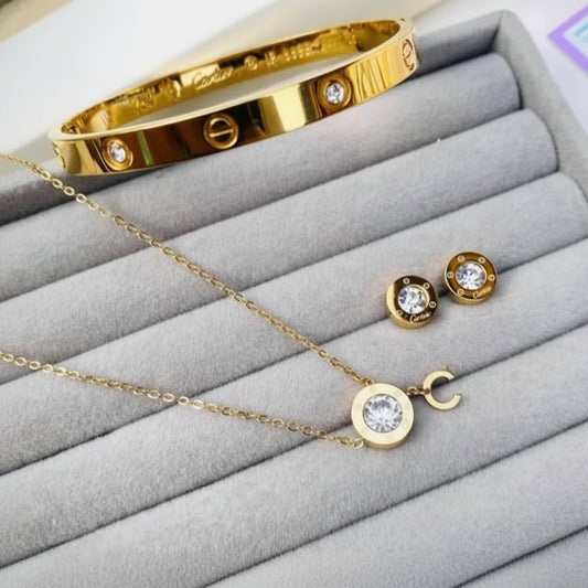 Gold jewelry set including a bangle bracelet, necklace, and stud earrings displayed on a gray velvet jewelry box. The pieces feature round clear gemstones in bezel settings.