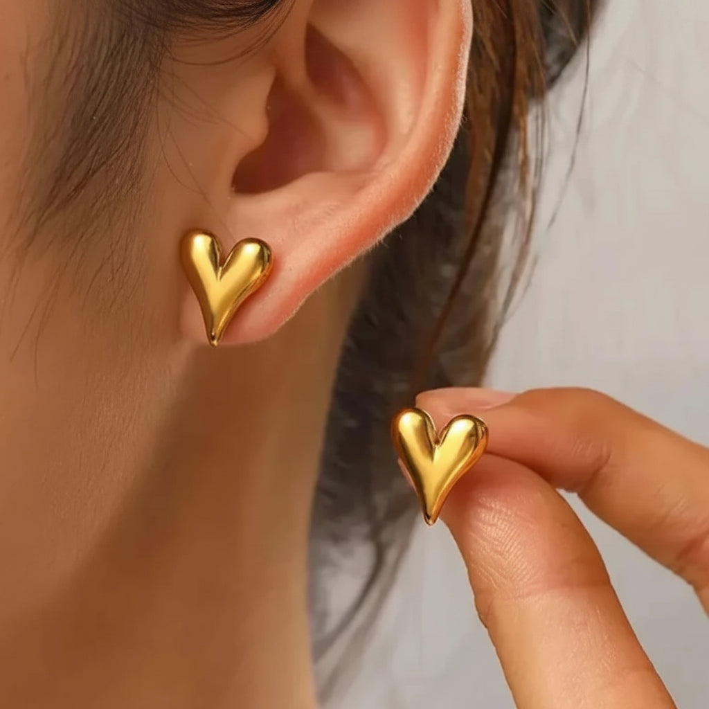 A close-up of a person wearing a small gold heart-shaped stud earring, with another hand holding a matching gold heart earring against a soft light gray background.