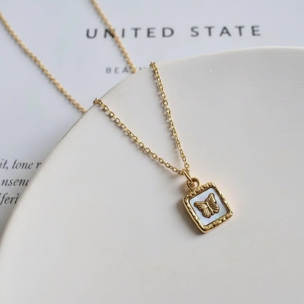 A gold chain necklace with a square pendant featuring a textured gold butterfly on a light gray inlay, displayed on a white surface.