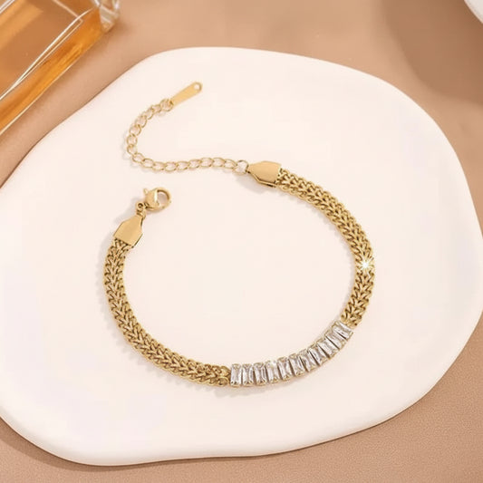A gold chain bracelet with a row of clear rectangular crystals across the front, displayed on a white ceramic dish against a beige background.