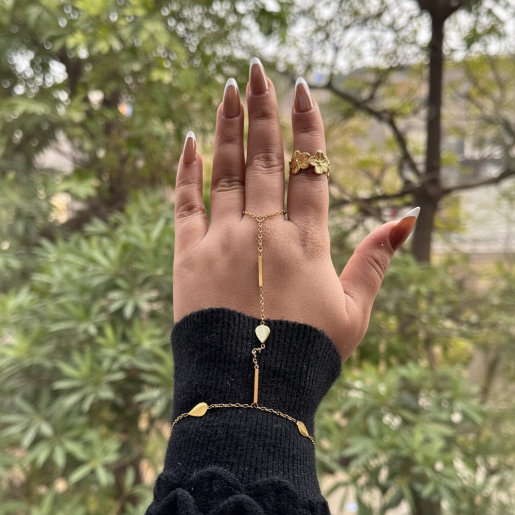 A gold chain hand bracelet with a central bar, small teardrop charms, and a heart-shaped pendant, shown worn on a person's hand with a black knit cuff, against a blurred outdoor background.