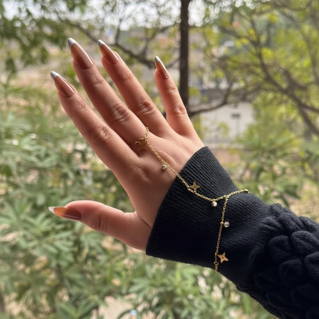 A gold star hand chain worn on a person's hand and wrist, paired with a black knit sweater sleeve, set against a blurred outdoor background of green foliage.