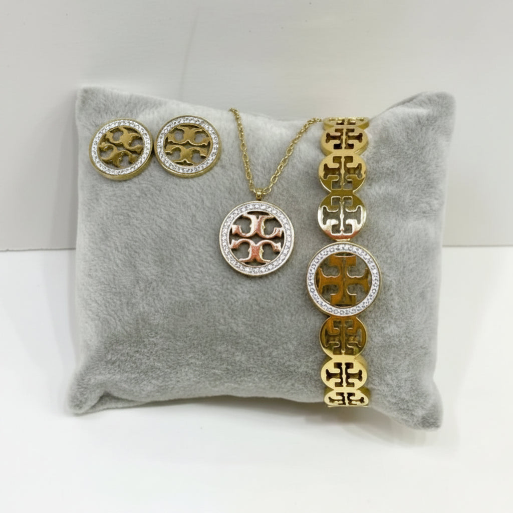 A gold-tone jewelry set including a necklace, bracelet, and matching earrings, displayed on a gray velvet cushion. The jewelry features a circular logo motif with orange enamel accents and clear rhinestone borders.