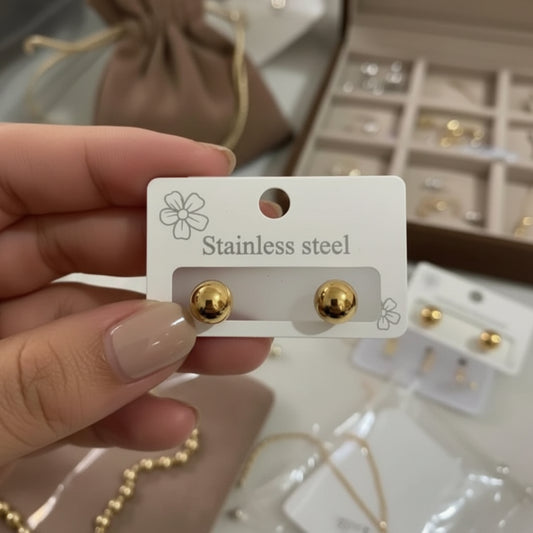 A hand holds a white card displaying a pair of small gold stud earrings labeled 'Stainless steel.' The background shows jewelry display boxes and additional earrings, with a soft-focus beige jewelry pouch nearby.