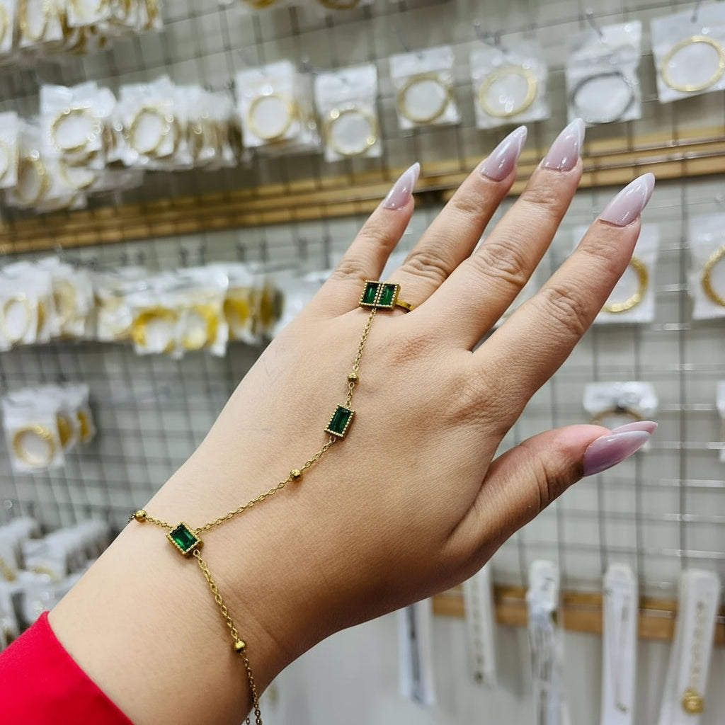 A gold chain bracelet and matching ring with square green stones, shown worn on a person's wrist and finger. The hand has long, manicured nails, and the background shows a retail display of other jewelry.