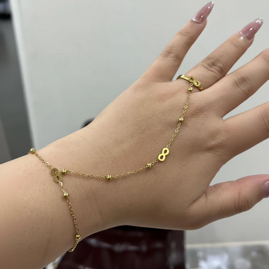 A delicate gold chain bracelet draped across a person's hand, featuring small gold beads and a small infinity symbol charm. A matching gold ring is also visible on the finger.