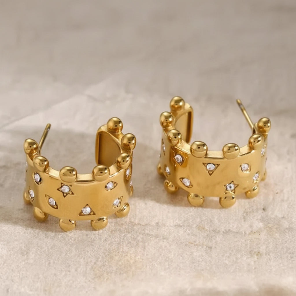 A pair of gold hoop earrings with a crown-like design, featuring small clear rhinestones set into the front, displayed on a textured light beige surface.