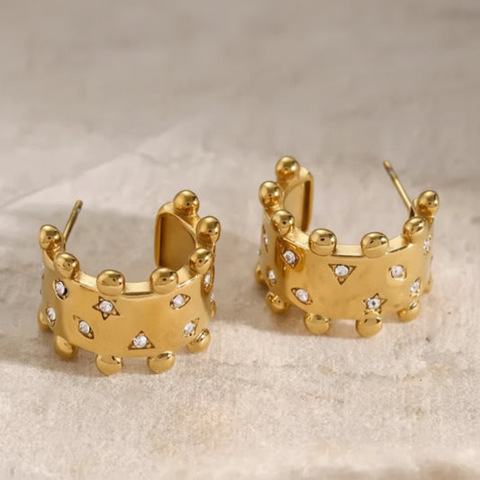 A pair of gold hoop earrings with a crown-like design, featuring small clear rhinestones set into the front, displayed on a textured light beige surface.