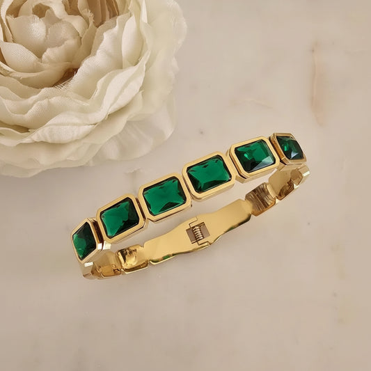 A gold-plated bracelet featuring seven square-cut emerald green rhinestones, displayed on a light beige marble surface next to a white fabric flower.