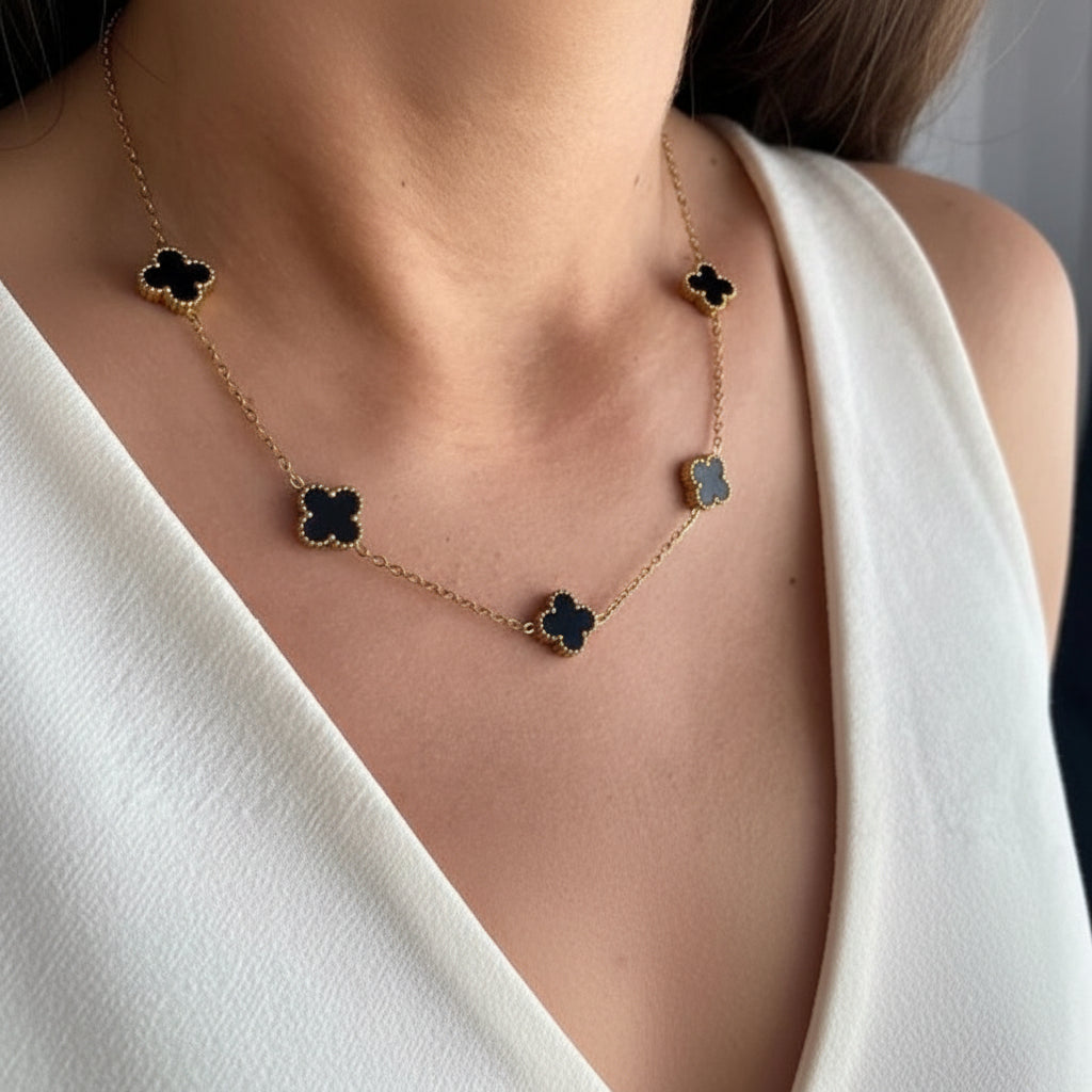 A gold chain necklace with five black clover motifs, worn by a model in a white sleeveless top.