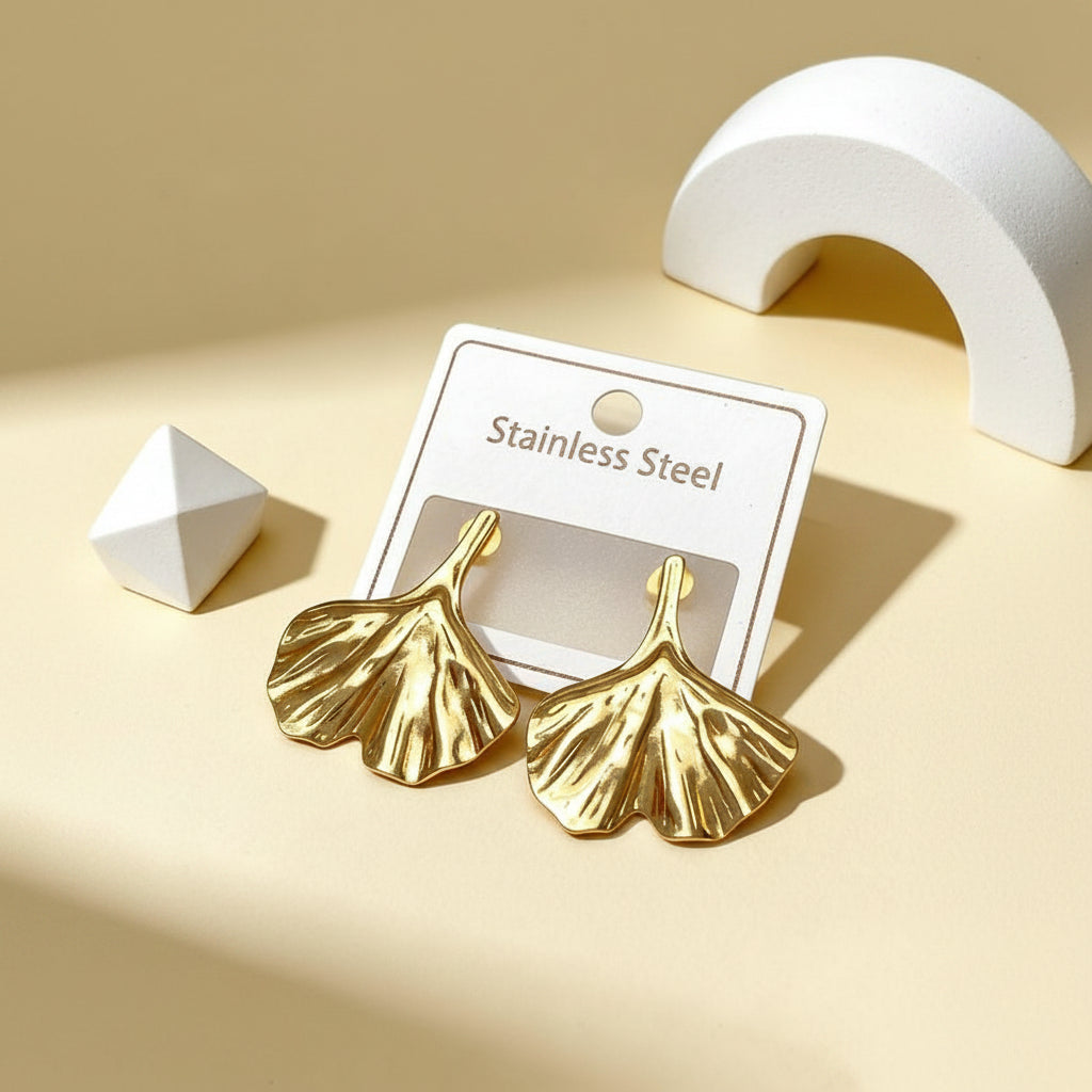 Gold leaf-shaped earrings on a white card with 'Stainless Steel' text, against a beige background.