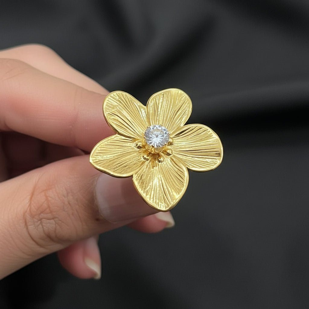 A gold flower-shaped ring with a textured petal design and a clear faceted gemstone in the center, held between fingers against a dark background.