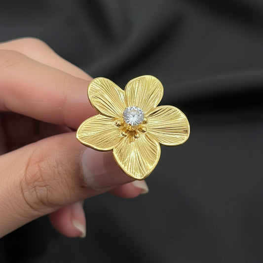 A gold flower-shaped ring with a textured petal design and a clear faceted gemstone in the center, held between fingers against a dark background.
