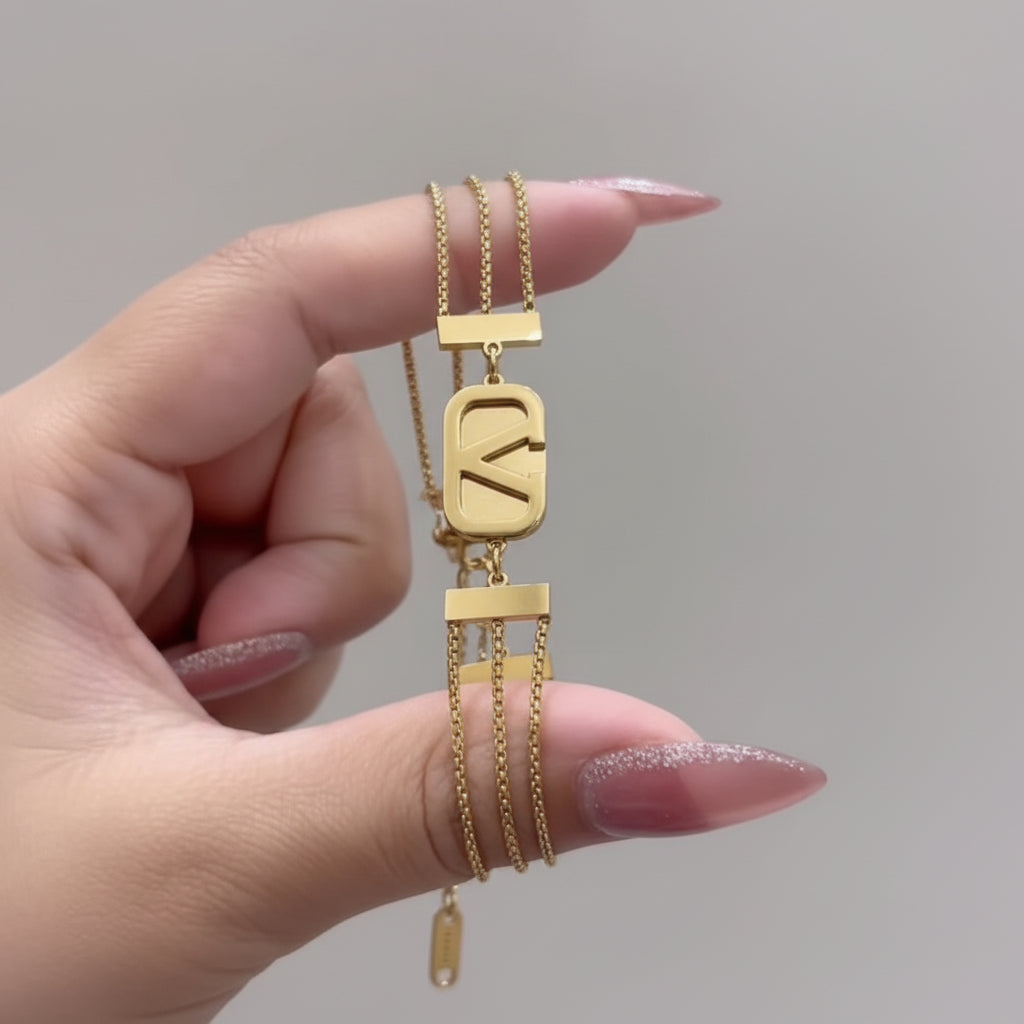 A close-up of a hand holding a gold multi-layered chain necklace with a large rectangular pendant featuring a stylized letter 'K' and smaller dangling chain elements.