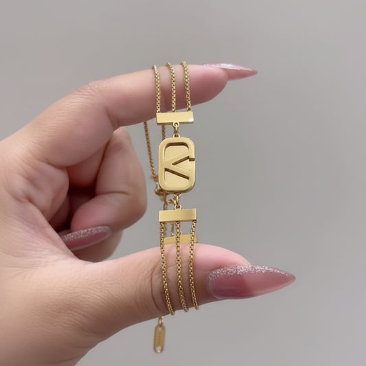 A close-up of a hand holding a gold multi-layered chain necklace with a large rectangular pendant featuring a stylized letter 'K' and smaller dangling chain elements.
