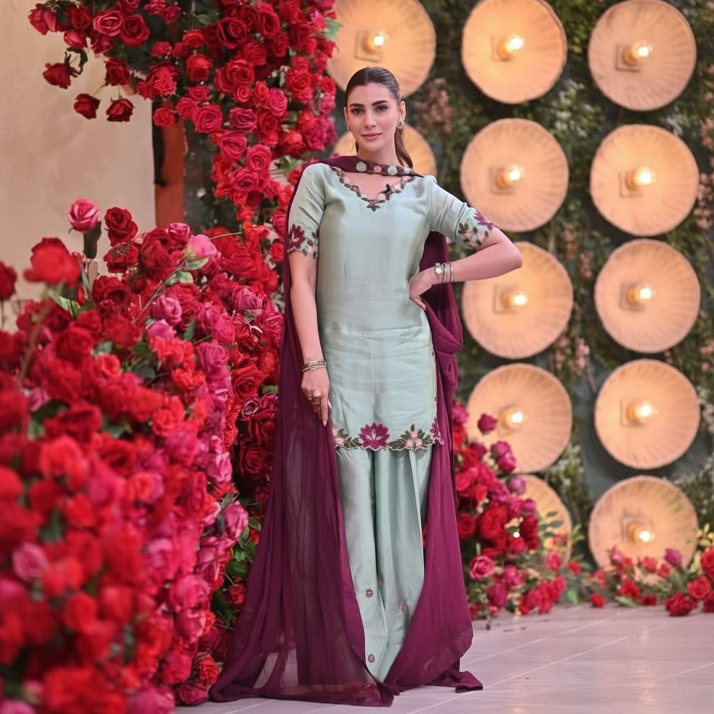 A woman modeling a light sage green traditional salwar kameez suit with a deep purple dupatta, standing in front of a decorative background of red roses and circular wooden wall panels.