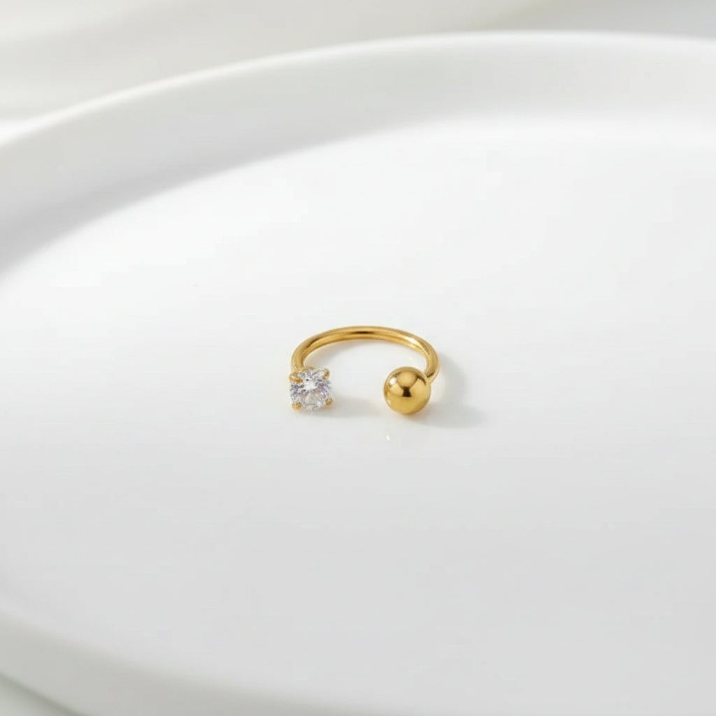 A small gold hoop-style body jewelry piece with a clear faceted gemstone and a polished gold ball end, photographed on a soft white fabric surface.