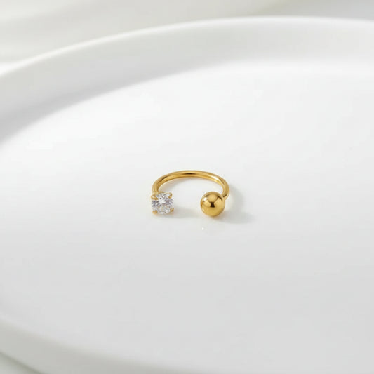 A small gold hoop-style body jewelry piece with a clear faceted gemstone and a polished gold ball end, photographed on a soft white fabric surface.