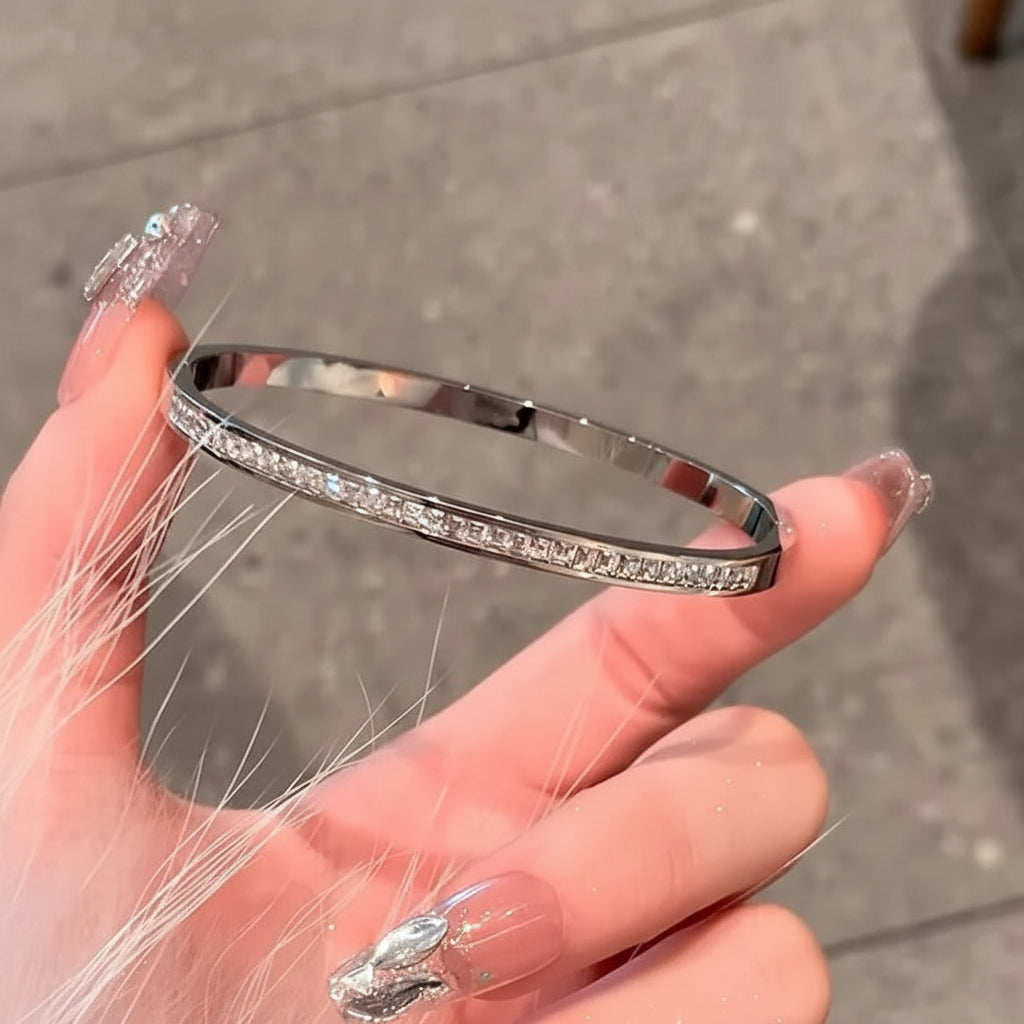 A polished silver-toned bangle bracelet with a row of clear stones along the front, held by a hand with decorated nails against a gray tiled background.