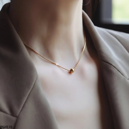 A close-up of a delicate gold chain necklace with a small spherical bead pendant, worn by a person in a taupe blazer.