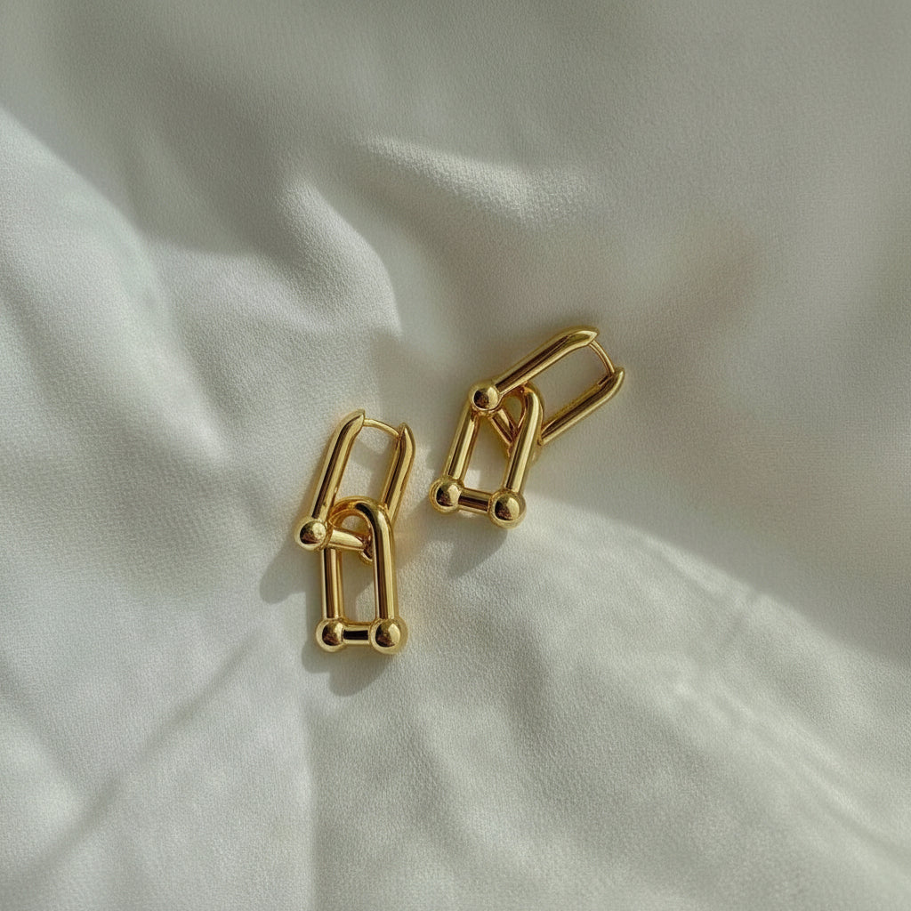A pair of gold-toned geometric hoop earrings with small bead accents, displayed on a softly textured white fabric background.