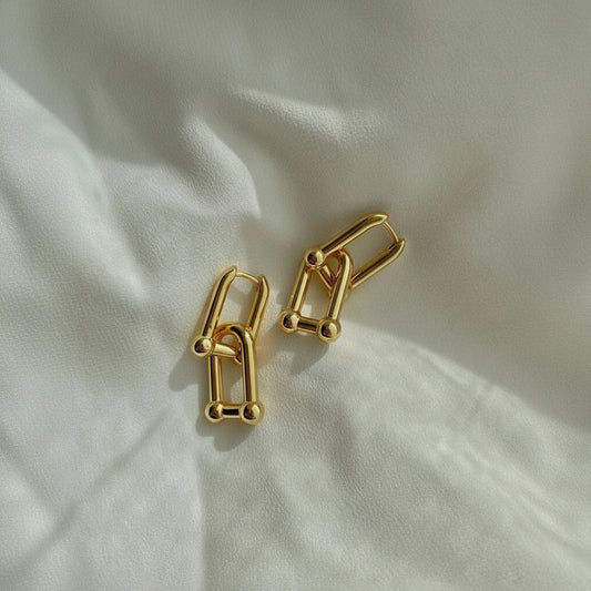 A pair of gold-toned geometric hoop earrings with small bead accents, displayed on a softly textured white fabric background.
