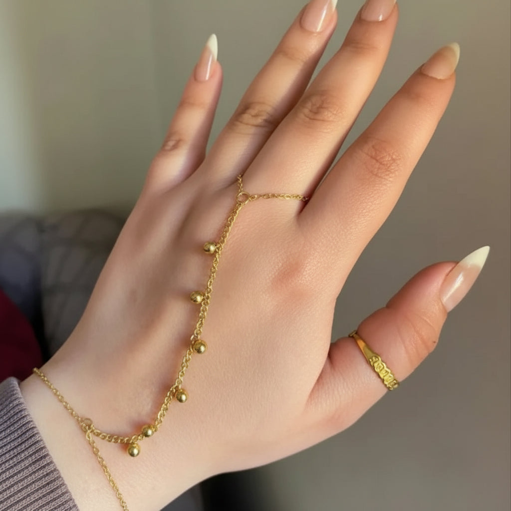A close-up of a hand wearing a delicate gold chain hand bracelet with small bead drops and a matching gold ring on the middle finger, against a soft beige background.