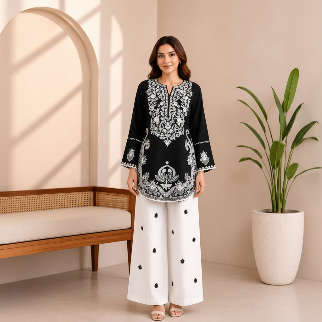 A woman modeling a black kurta with intricate silver embroidery and white wide-leg pants with black polka dots, standing in a brightly lit, modern room with a wooden bench and a potted plant.