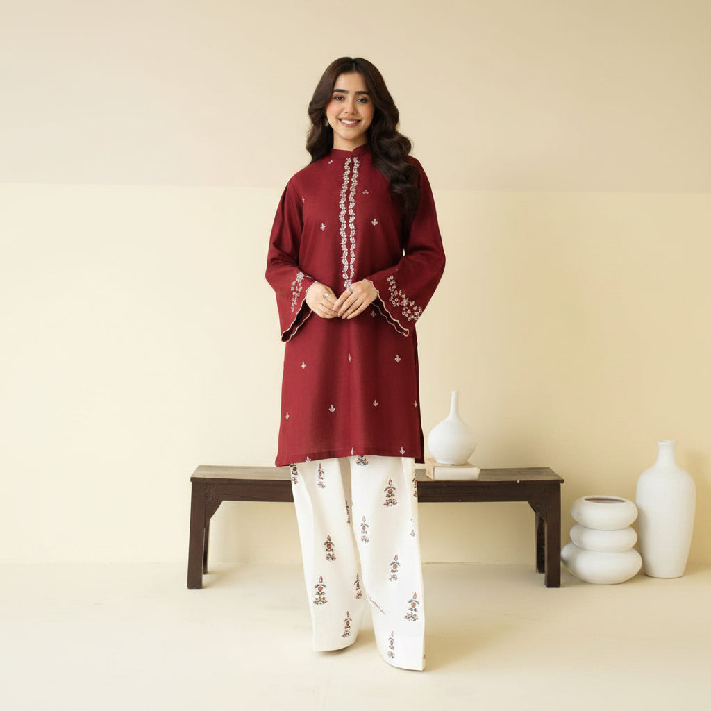 Woman wearing a maroon embroidered top and white pants against a beige background