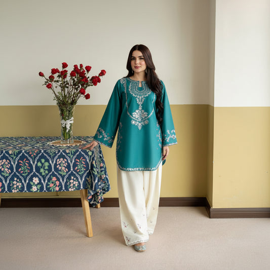 Woman in teal embroidered kurta with white pants standing in a room with a table and vase of flowers.