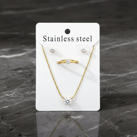 A jewelry set displayed on a white backing card labeled 'Stainless steel,' including a gold-tone ring with a clear stone, a gold-tone necklace with a round clear stone pendant, and matching stud earrings. The card is photographed on soft white fabric.
