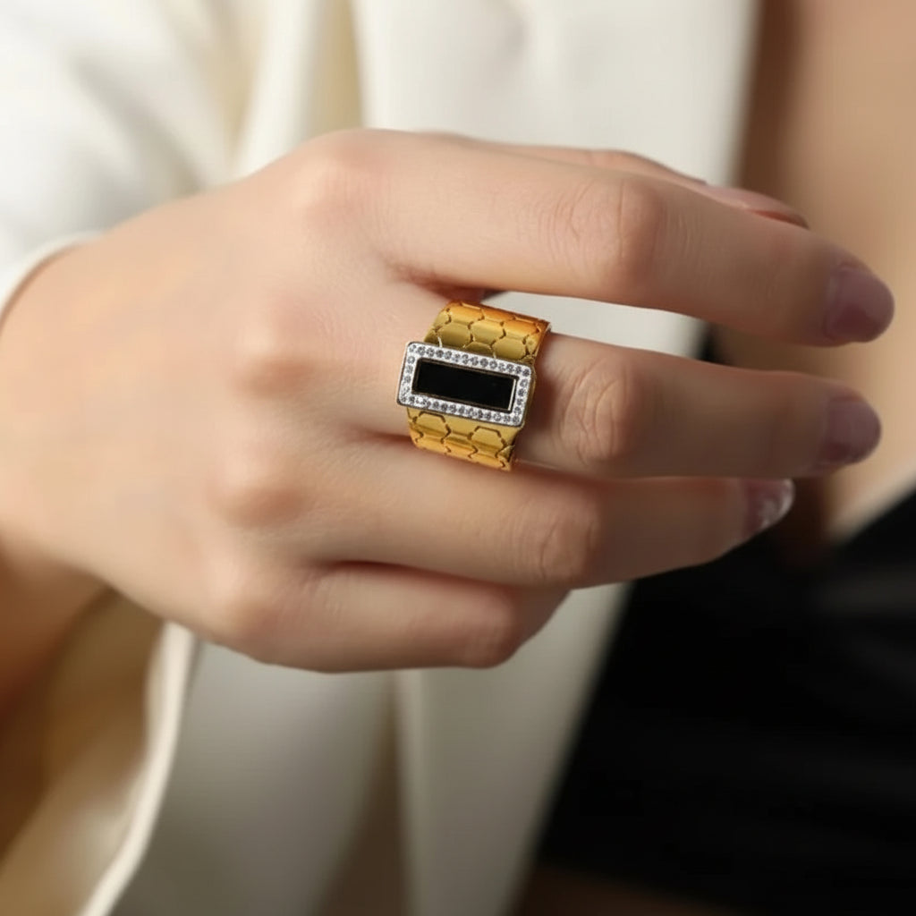 A close-up of a hand wearing a statement ring. The ring features a textured gold band with a central black rectangular inlay, bordered by a halo of small clear stones on the top and sides.