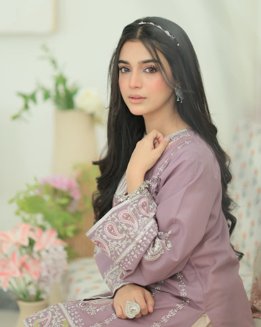 A woman wearing a lavender traditional outfit with intricate silver embroidery and paisley patterns, posed in a softly lit indoor setting with blurred flowers in the background.