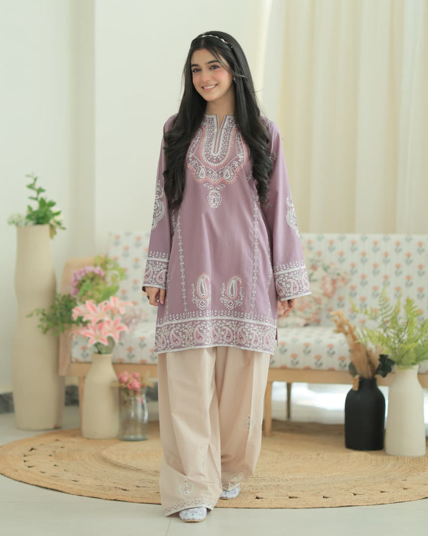 A young girl modeling a traditional three-piece salwar kameez suit in a bright, modern indoor setting. The set features a lavender purple embroidered kurta with beige wide-leg trousers and a matching light beige top layer.