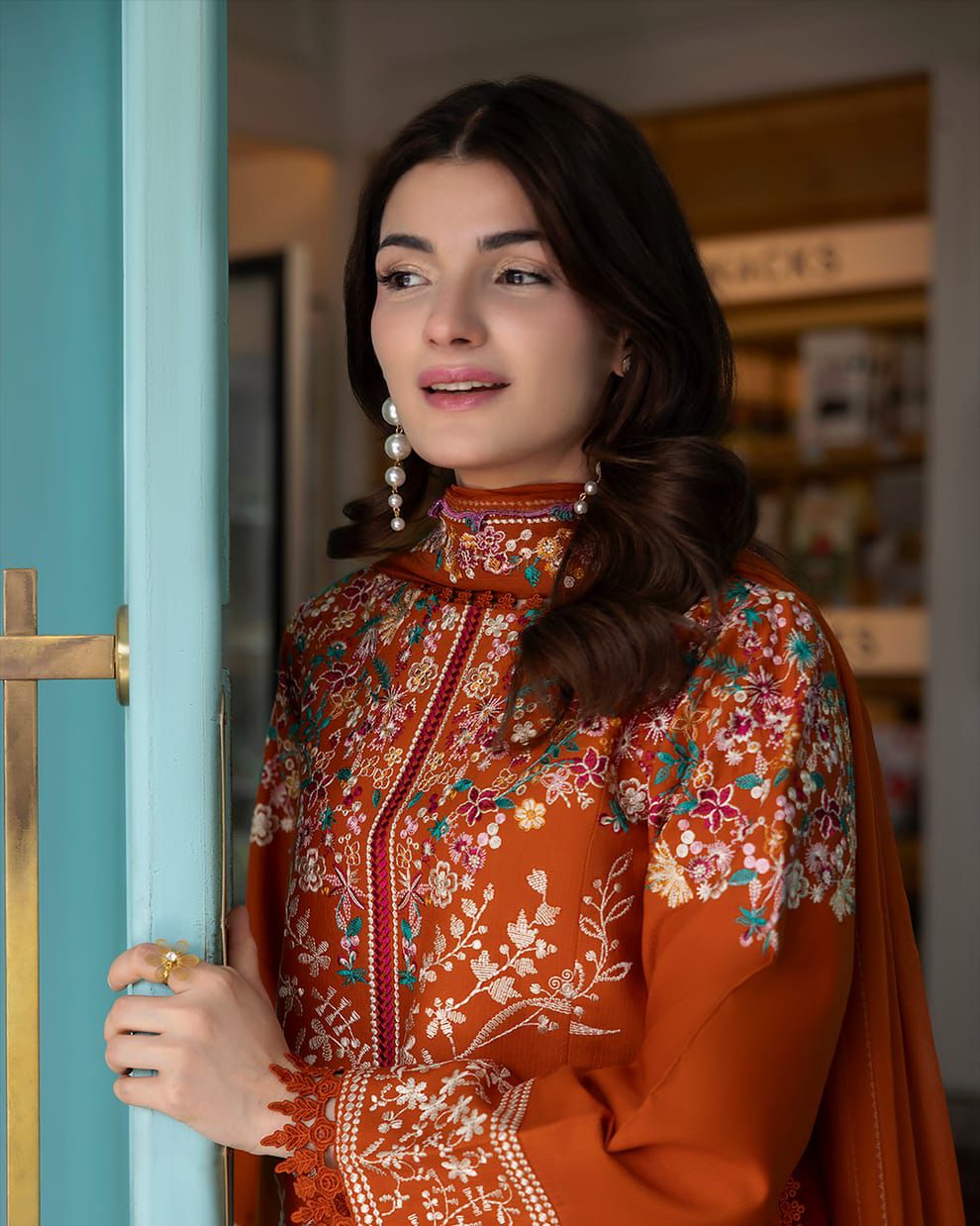 A woman wearing a rust-orange embroidered traditional outfit with floral patterns and a high collar, standing next to a turquoise door.