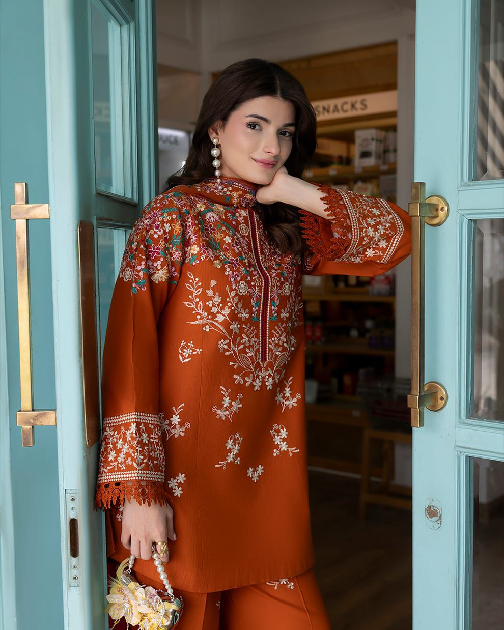 A woman models an orange traditional embroidered kurta and matching trousers, leaning against a turquoise door in a shop setting.