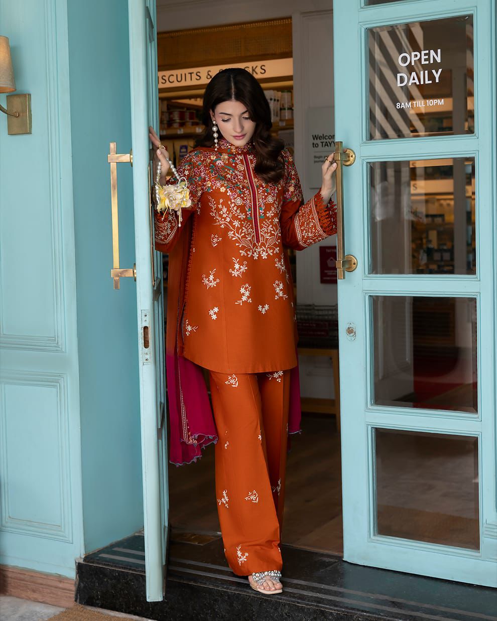 A woman models an orange rust-colored traditional South Asian outfit with floral embroidery, standing in a doorway of a light blue shop.