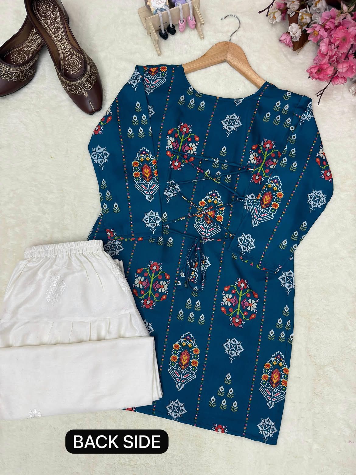A dark teal printed kurta with multicolor floral and geometric patterns, shown on a wooden hanger next to folded white trousers, with a 'BACK SIDE' label at the bottom.