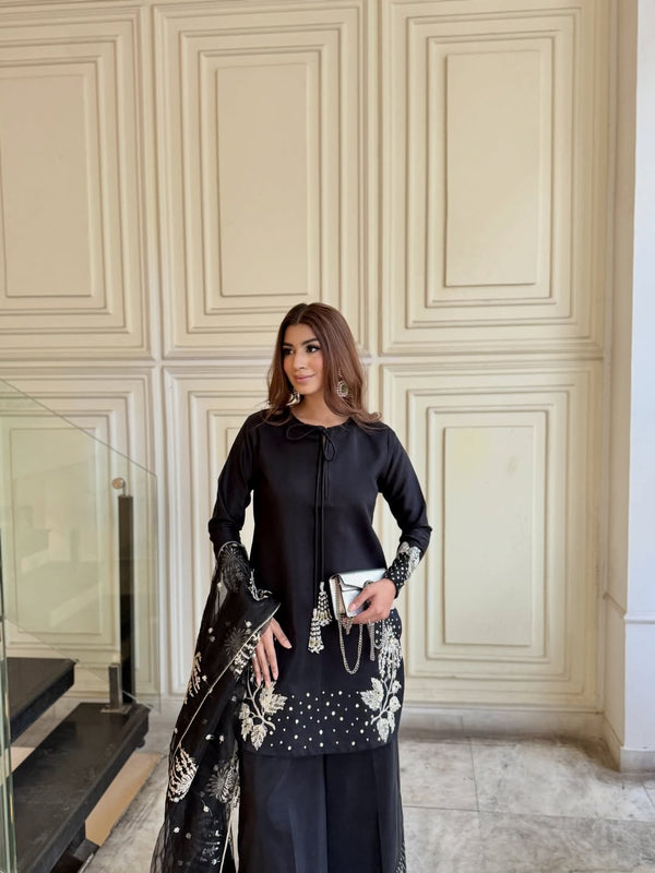 A woman models a black traditional outfit with intricate silver and white embroidery, featuring a long-sleeve top, matching pants, and a patterned dupatta, standing in front of a cream-colored paneled wall.
