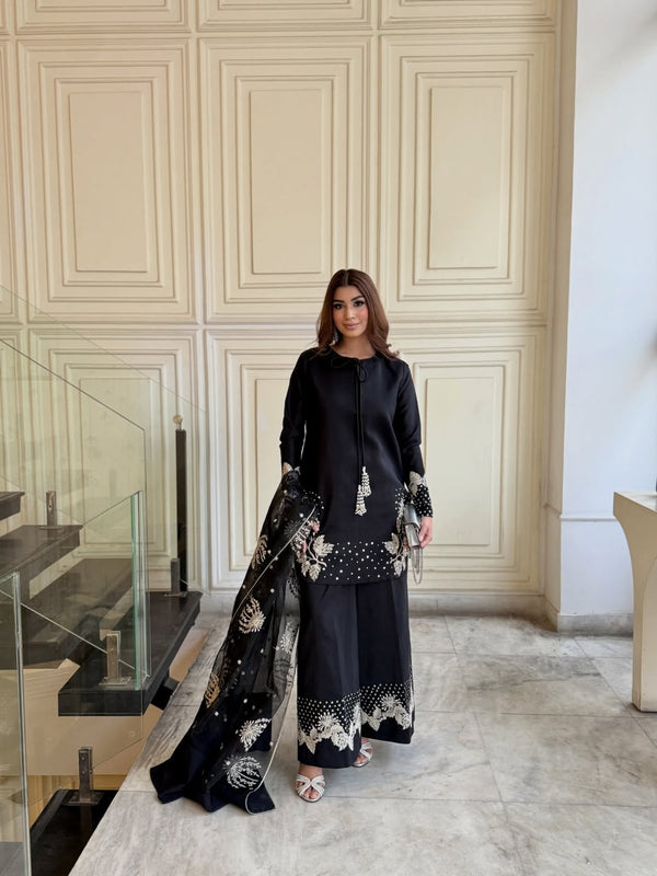 Woman in a black outfit with floral patterns standing in a room with white paneled walls.
