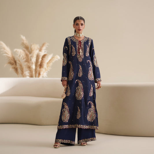 Woman wearing a navy blue embroidered outfit with gold patterns in a minimalistic setting.