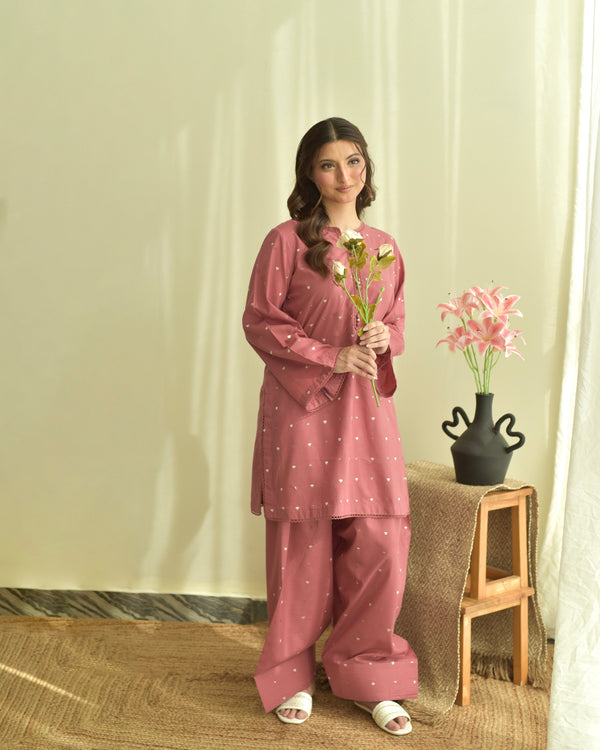 Woman in a pink traditional outfit holding flowers in a room with a vase on a stool.