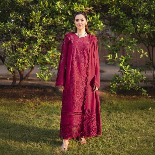 A woman stands outdoors on grass in front of leafy trees, wearing a long maroon embroidered dress with matching draped sleeves and a sheer embroidered overlay.