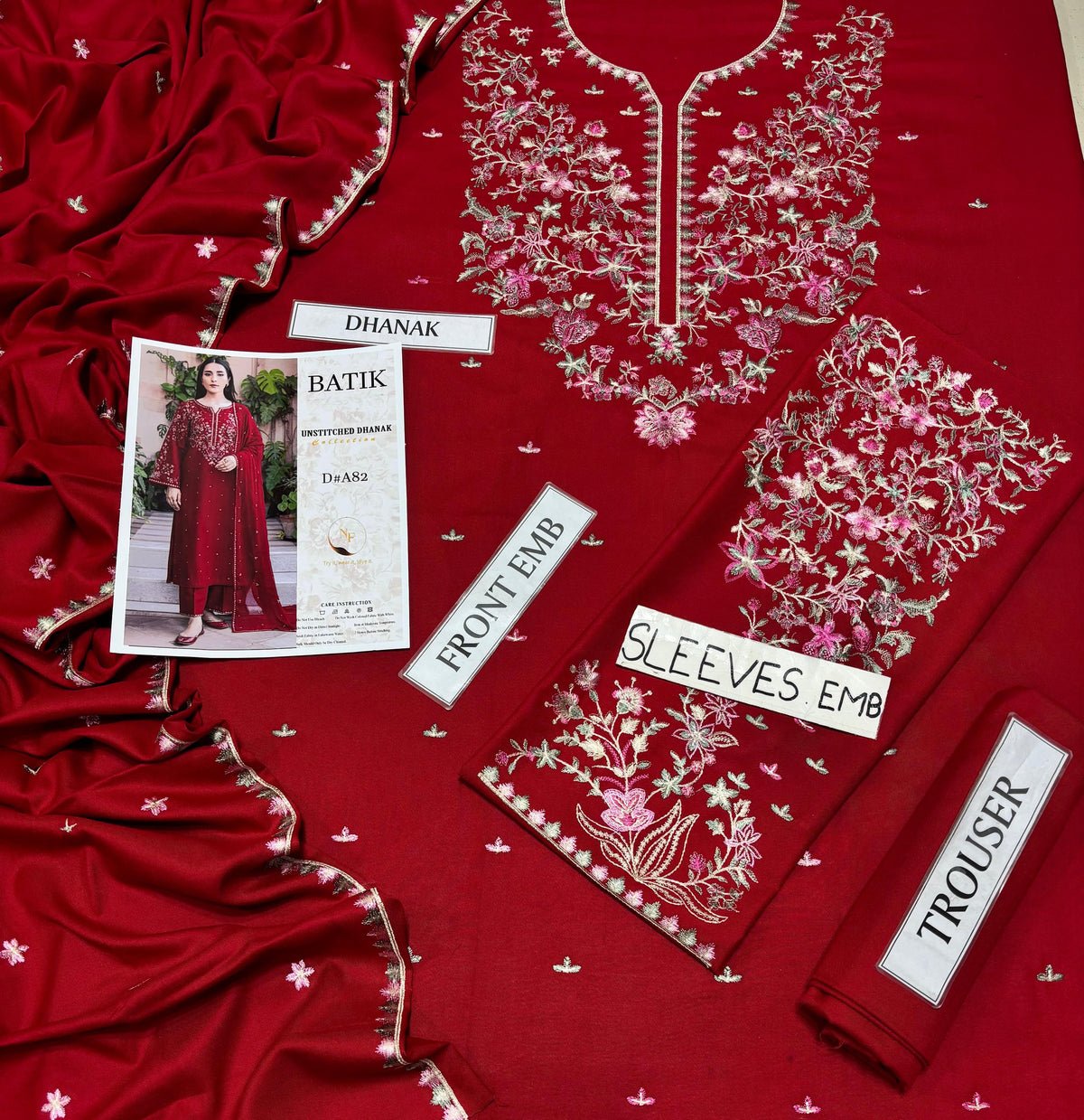A red embroidered women's suit set laid flat on a white background, including a kurta front panel, sleeves, trouser, matching fabric, and a printed card showing a model wearing the outfit. White labels identify parts such as FRONT EMB, SLEEVES EMB, and TROUSER.